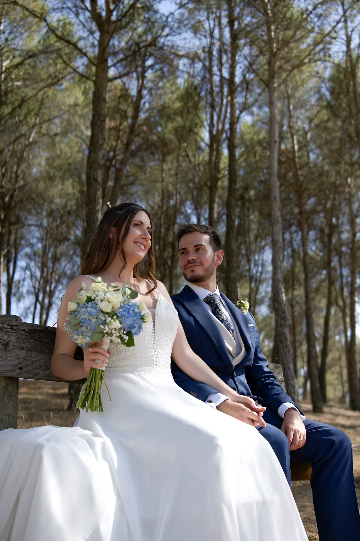 Raquel y Jes&uacute;s sentados en un banco bajo los pinos, ella con el ramo