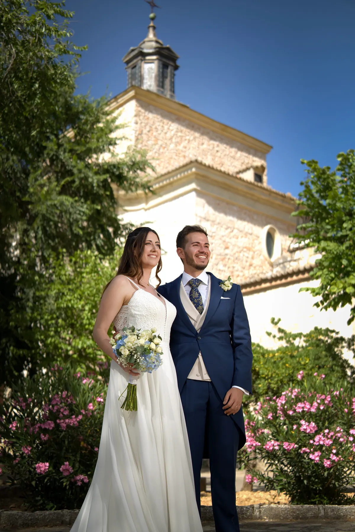 Jes&uacute;s y Raquel, reci&eacute;n casados, ante la fachada de la iglesia entre adelfas